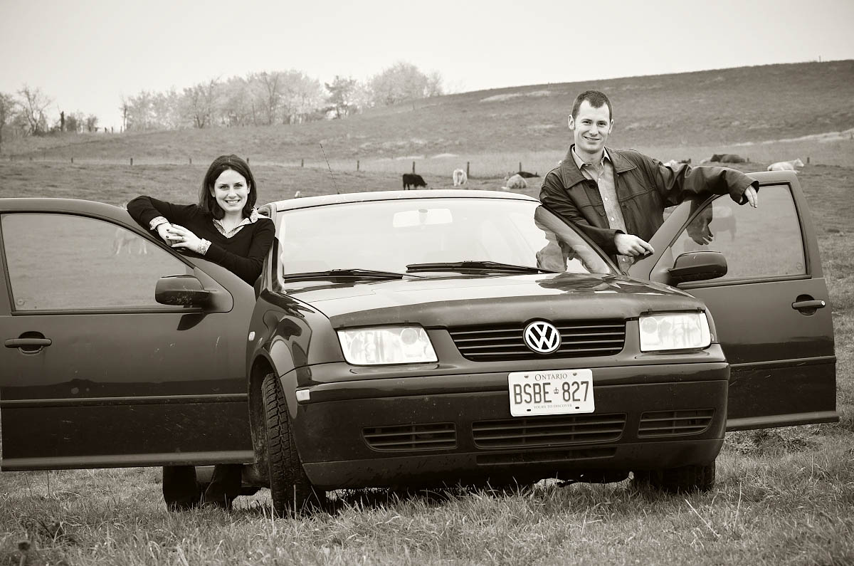 Young couple pose with their first car in farm field