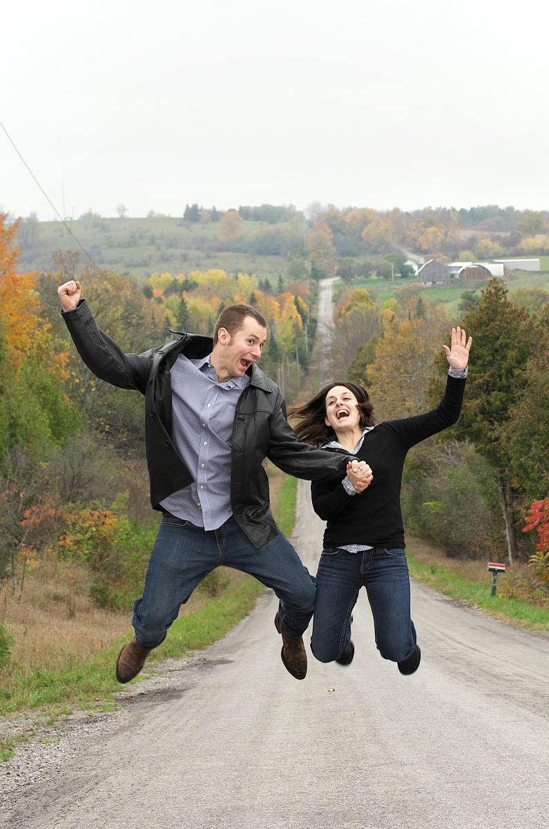 Couple jumps for joy on steep country road for country themed Peterborough engagement photography session