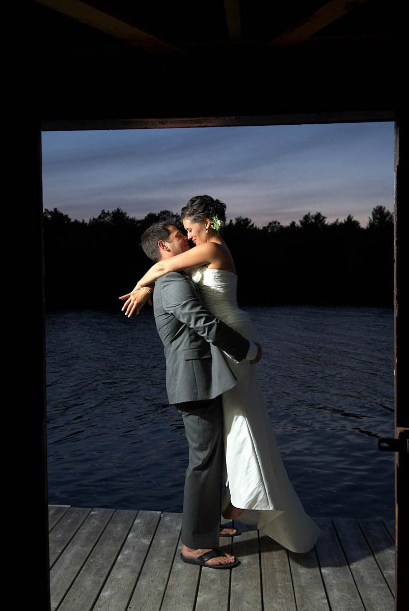 At dusk, groom lifts his bride in his arms outside boathose on Stoney Lake