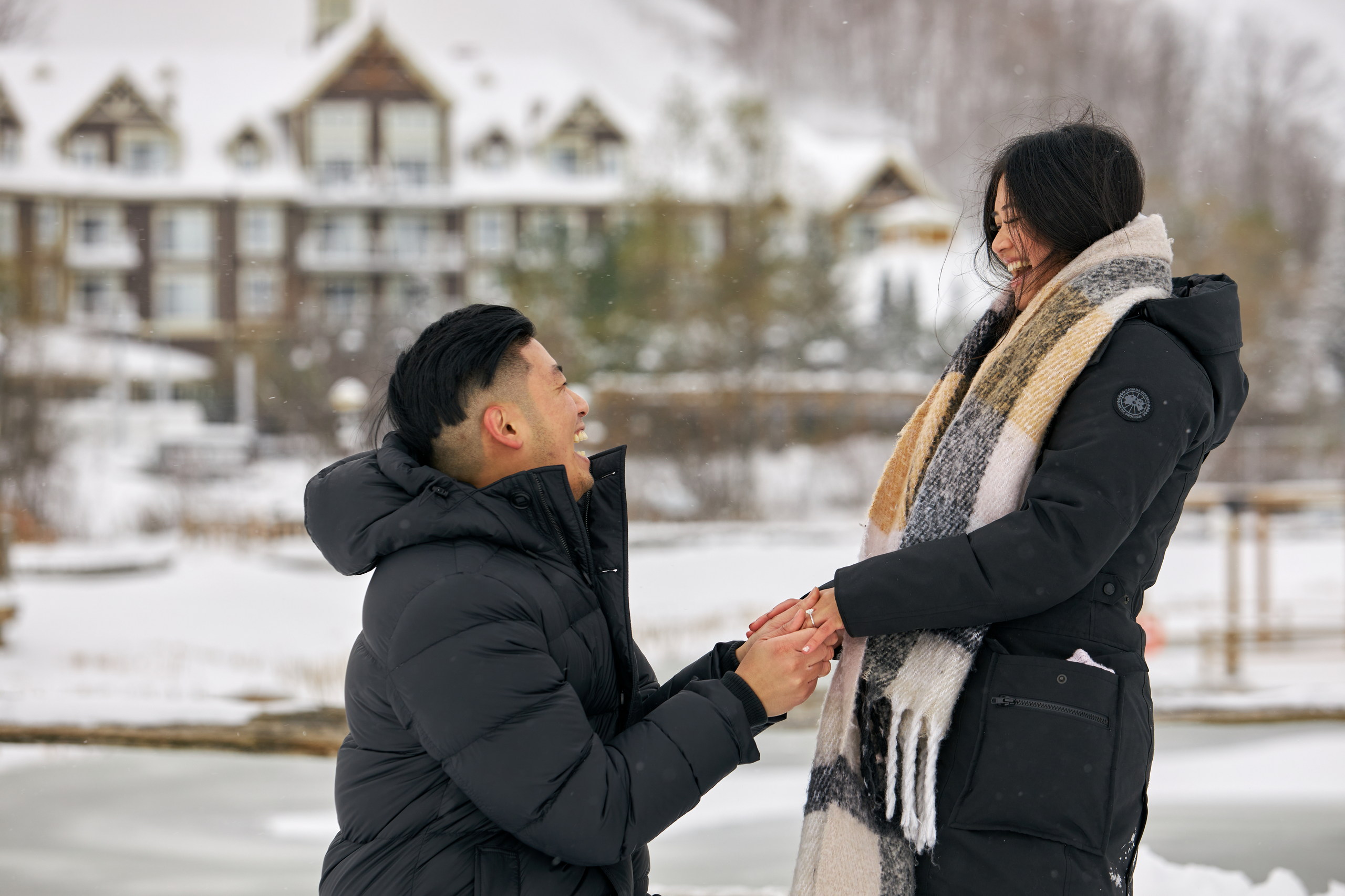 A young man on bent knee asks his girlfriend to marry him in front of Millpond at Blue Mountain Village in winter